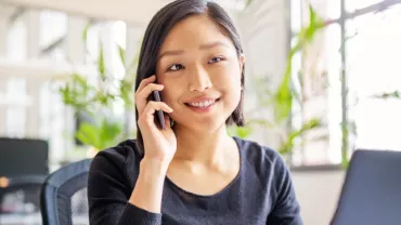 female Chinese white-collar professional in her 30s answering her mobile phone while sitting in front of a laptop in an office setting.